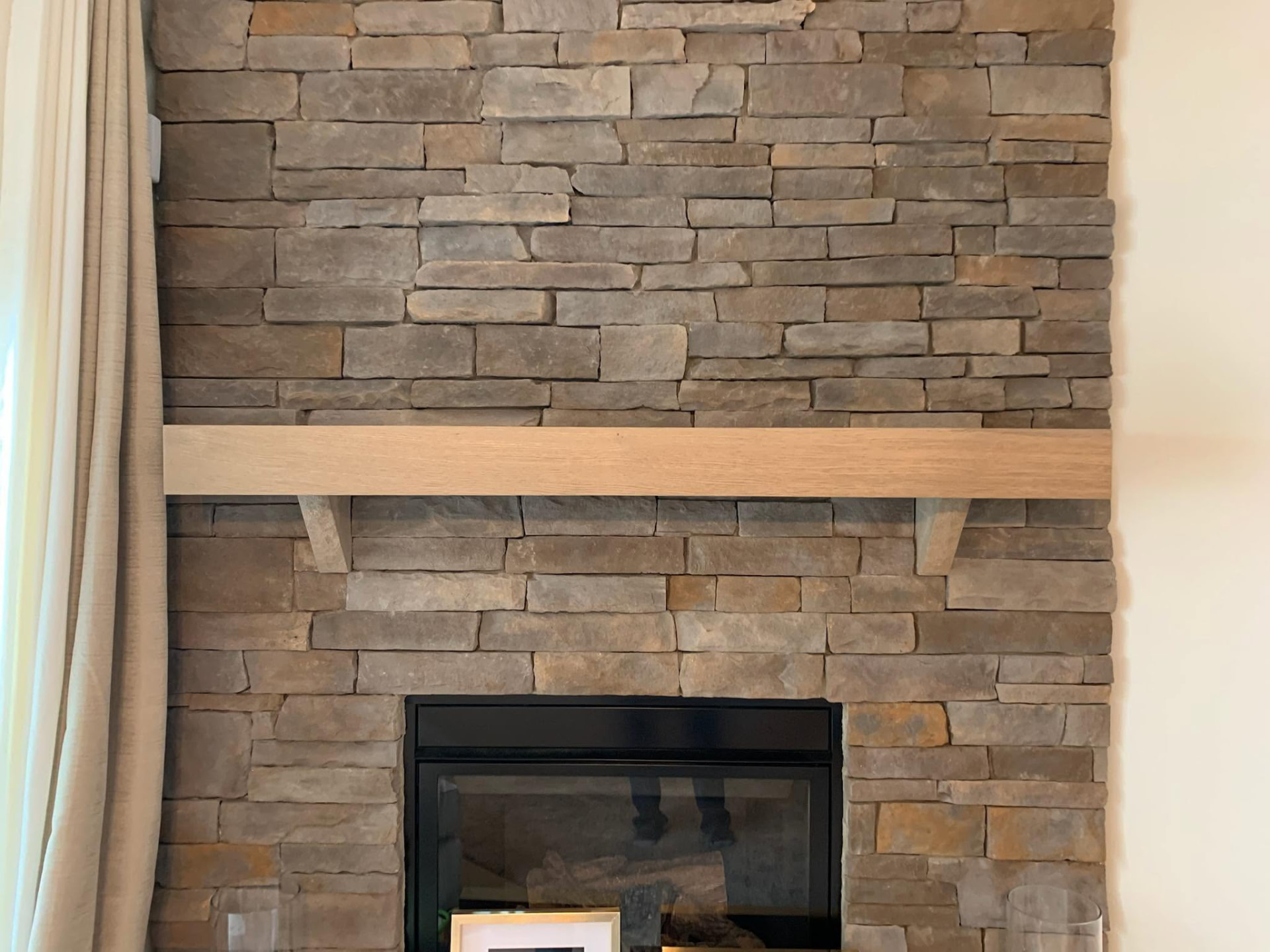 Thick pale white oak mantel beam mounted on a grey stacked stone fireplace
