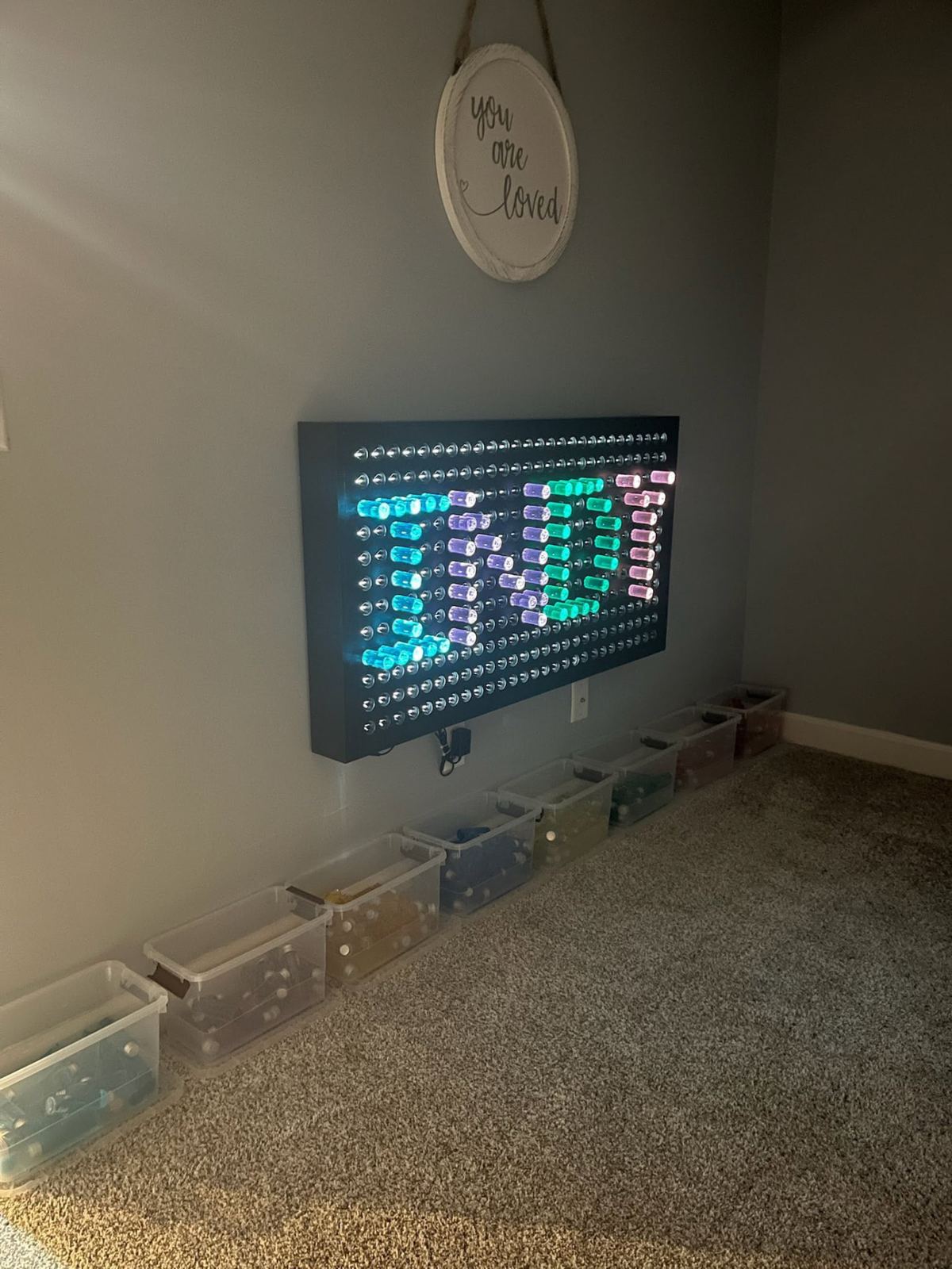 Dark wall-mounted plywood LED peg board light wall with colored pegs and sorting bins below